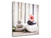 O imprimare pe pânză cu pietre de spa gri și negre, o floare roz de gerbera și prosoape albe, pe un fundal de bambus, cu o ramă vizibilă din lemn, Papilora.