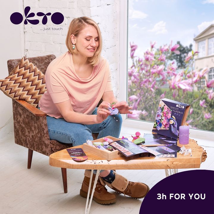 Un aranjament floral 3D plin de culoare, cu nuanțe de roz și violet, este expus într-un interior modern, deasupra unei console din lemn cu o vază și o lumânare, Papilora.